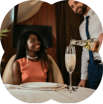 Mulher sentada com uma pessoa lhe servindo um champanhe em uma taça de vidro sobre a mesa.