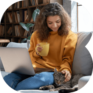 Mulher sentada em um sofá, enquanto faz carinho em seu gatinho de estimação, tomando seu chá ao mesmo tempo com seu notebook no colo.